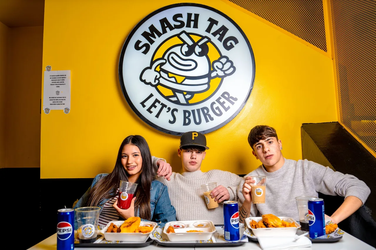 Ragazzi a tavola sotto il grande logo Smash Tag “Let’s burger”